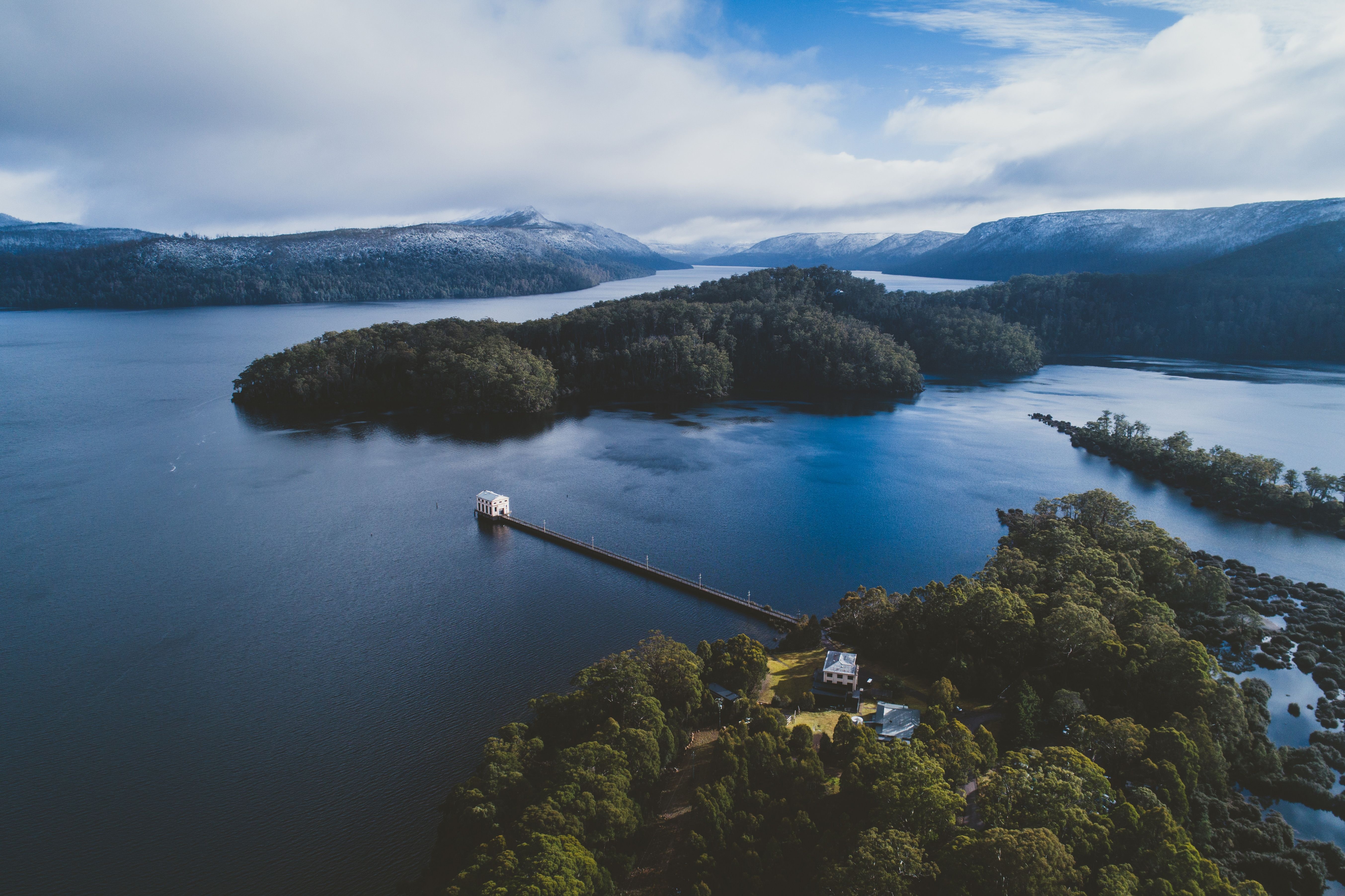 An aerial shot shows Pumphouse Point, a white building at the end of a long pier, extending into the dark blue waters of Lake St Clair. The pier connects to a densely forested landmass where other buildings are visible. In the background, snow-capped mountains and more forested islands rise from the expansive lake under a cloudy sky.