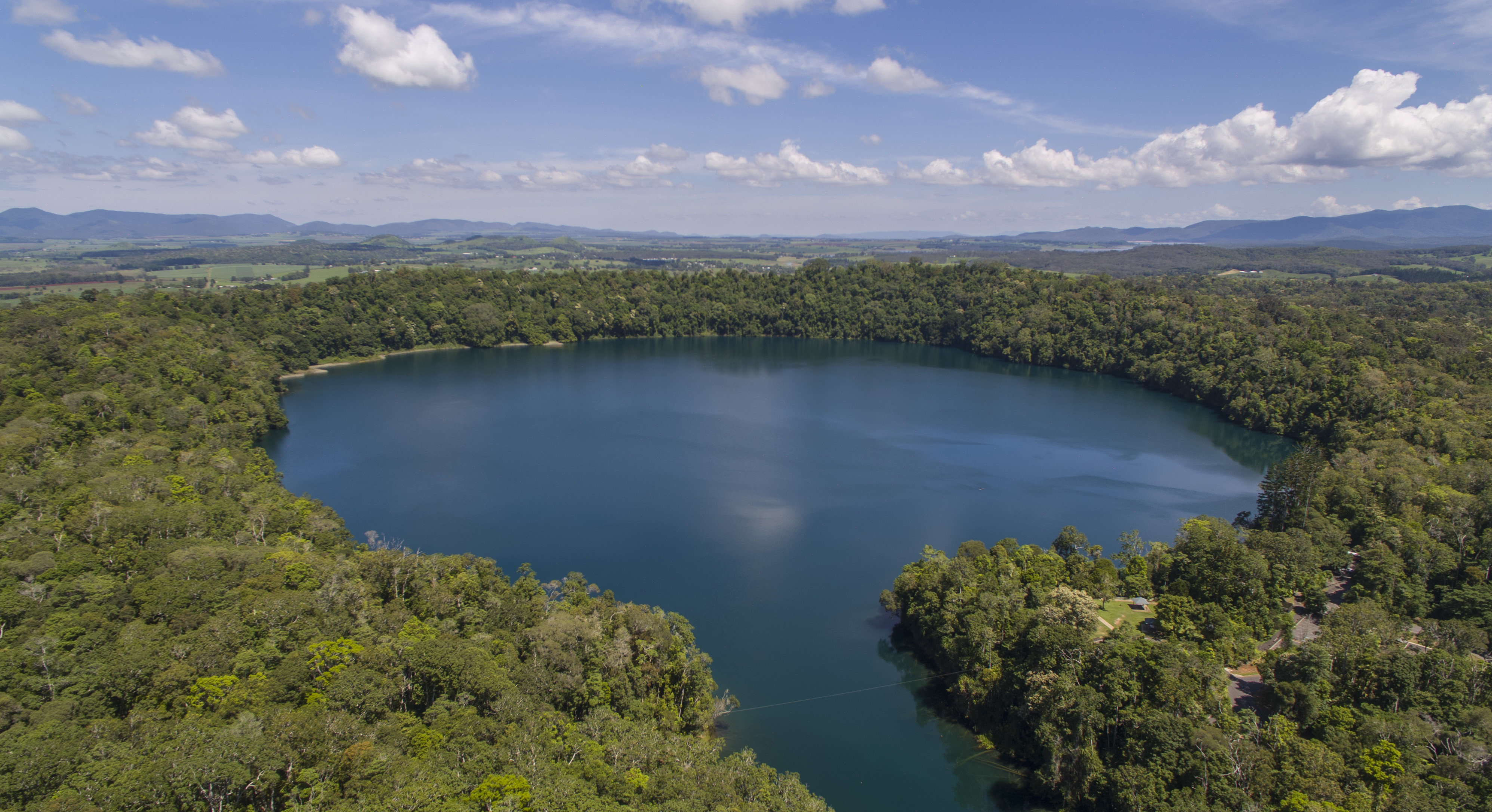 An aerial shot shows the perfectly circular, deep blue waters of Lake Eacham, a volcanic crater lake, completely encircled by lush, vibrant green tropical rainforest. Beyond the immediate tree line, agricultural fields and distant mountains are visible under a bright blue sky with scattered clouds.