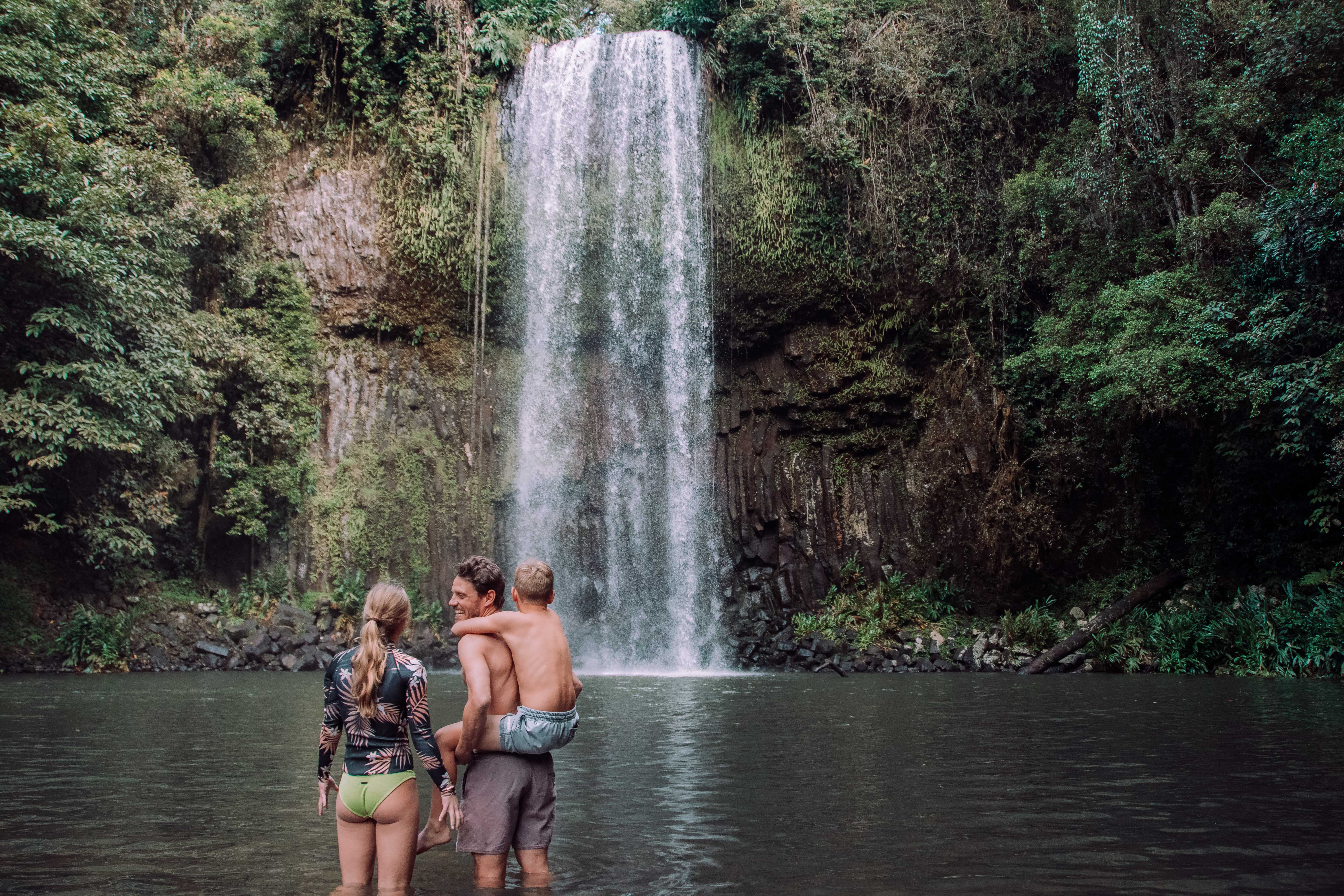 A man holding a child on his shoulders, standing in a natural pool with a woman beside him, all facing the majestic Millaa Millaa Falls cascading down a basalt cliff, surrounded by dense green rainforest.