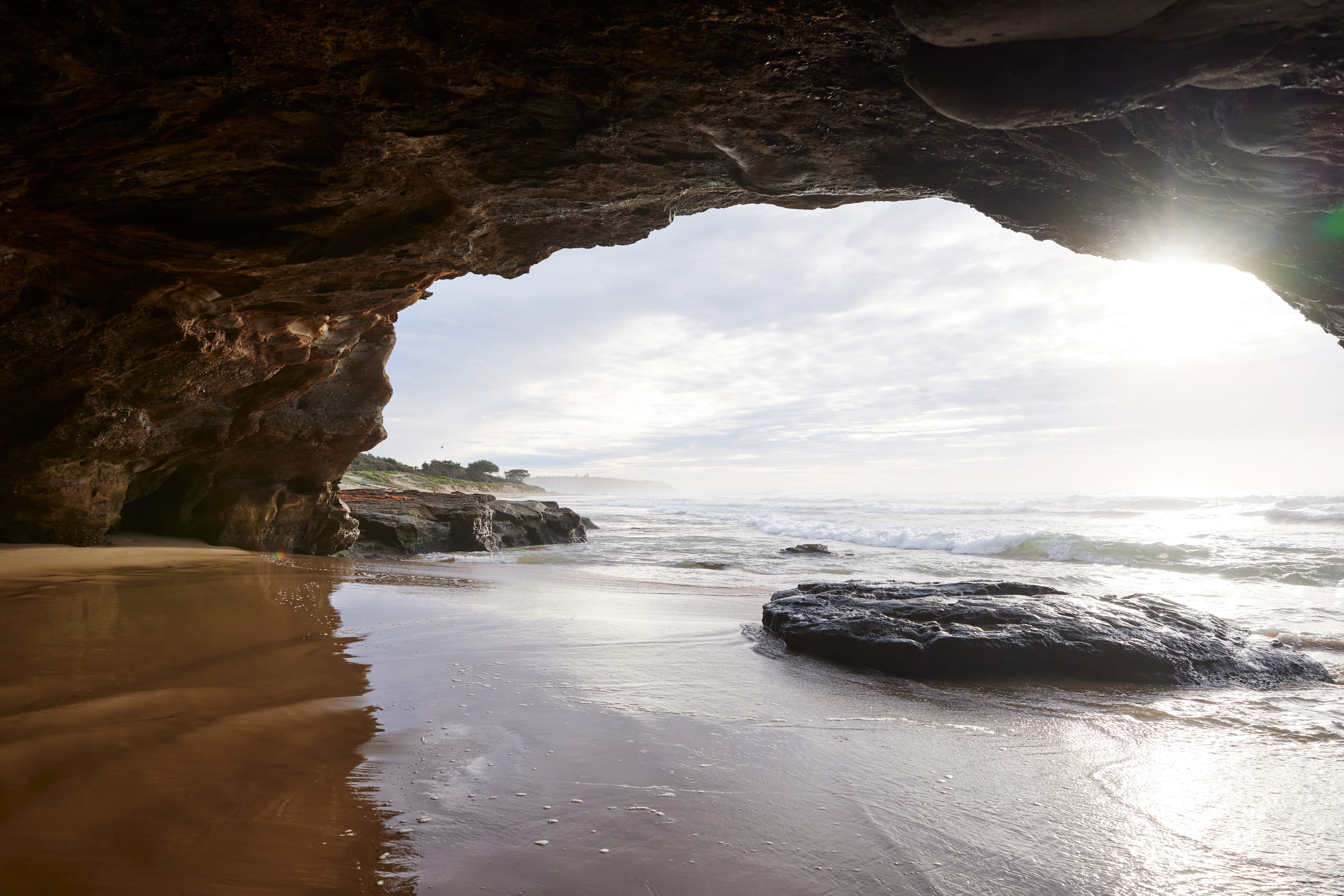 Looking out from the dark, textured entrance of a sea cave onto Caves Beach, Lake Macquarie. Sunlight streams from the upper right, illuminating the wet sand, a large dark rock, and white ocean waves crashing on the shore.