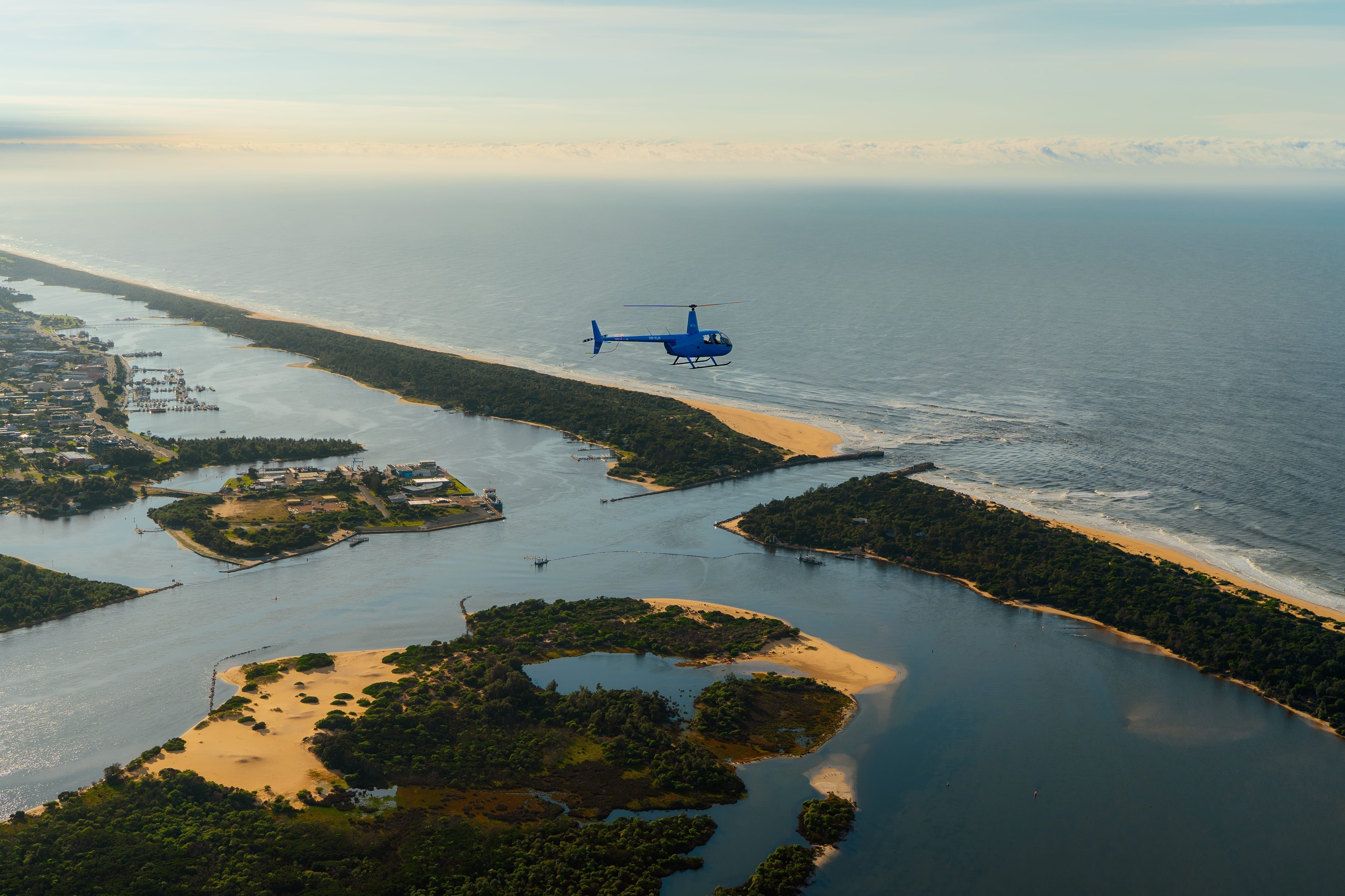 An aerial shot of Lakes Entrance, Victoria, featuring a blue helicopter flying above a long, narrow strip of land that separates a calmer inlet with boats and buildings from the open ocean. Sandy beaches and green vegetation line both sides of the land bridge.