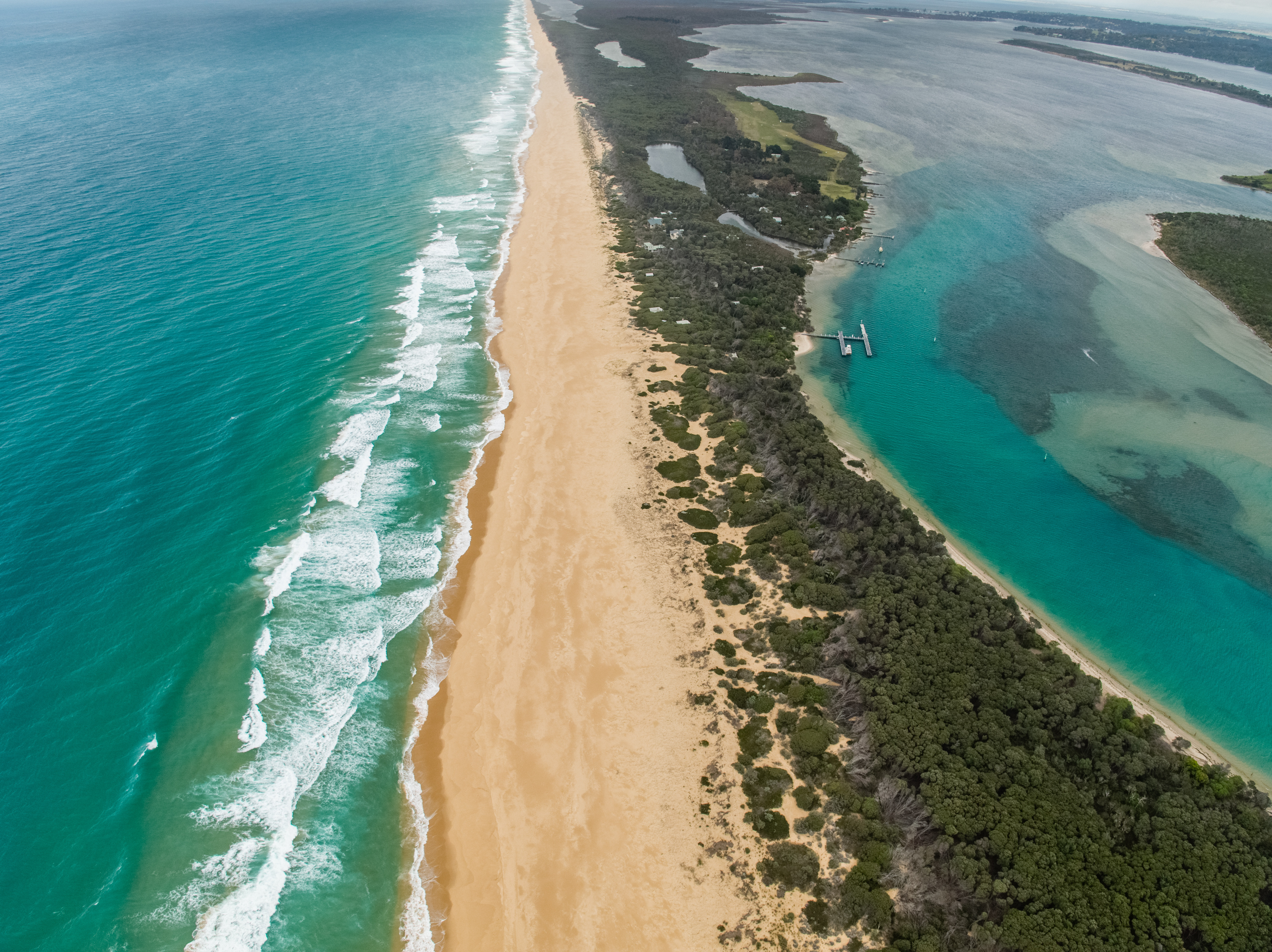 An aerial shot capturing the immense length of Ninety Mile Beach, with turquoise ocean waves breaking on a wide golden sand beach on the left, and a calm, darker blue inland waterway bordered by dense green vegetation on the right.