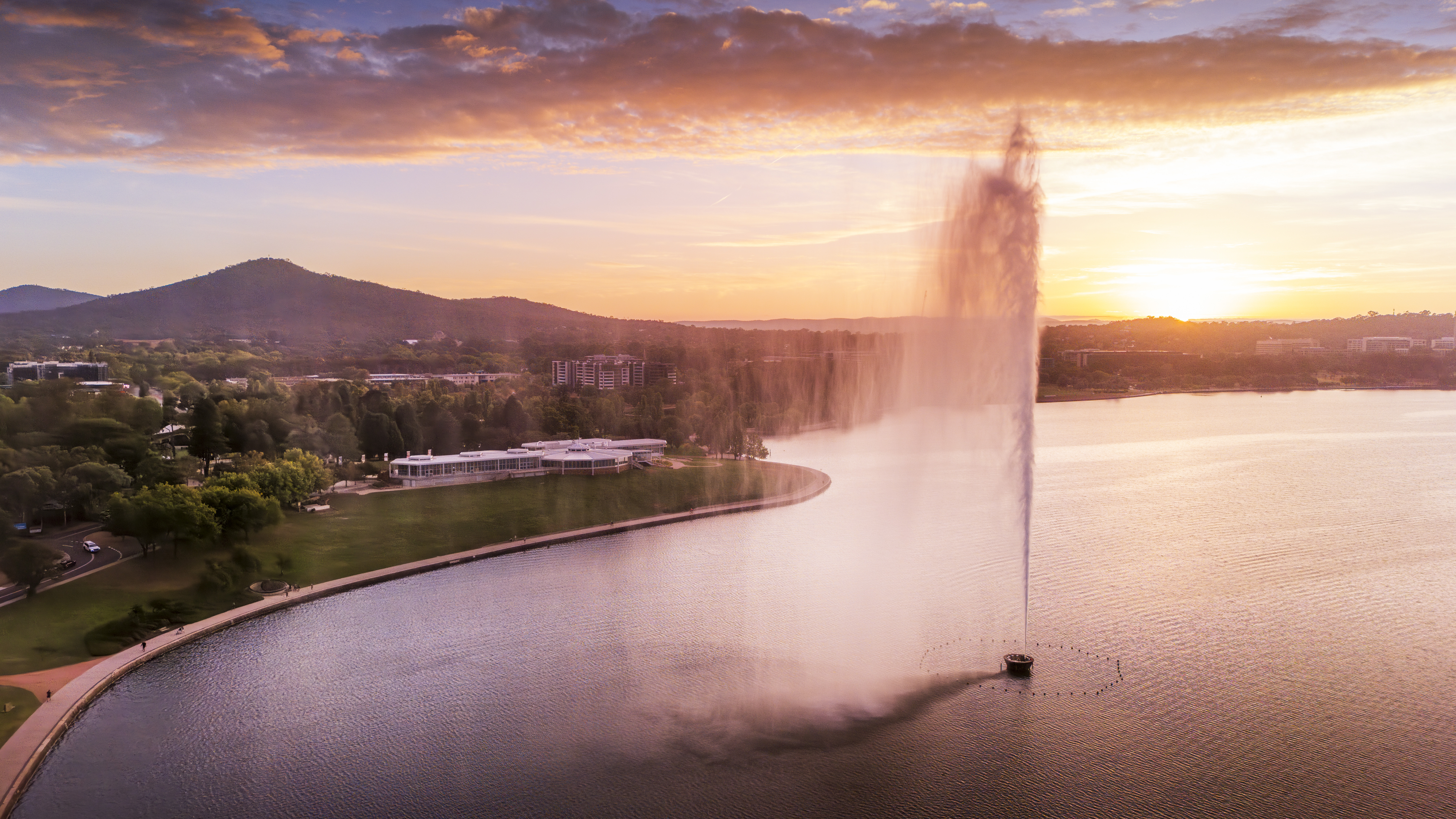 An aerial view of Lake Burley Griffin at sunset, with the powerful Captain Cook Memorial Jet sending a tall plume of water into the air. The golden and pink sky reflects on the lake, which is bordered by green parkland and distant buildings and hills.