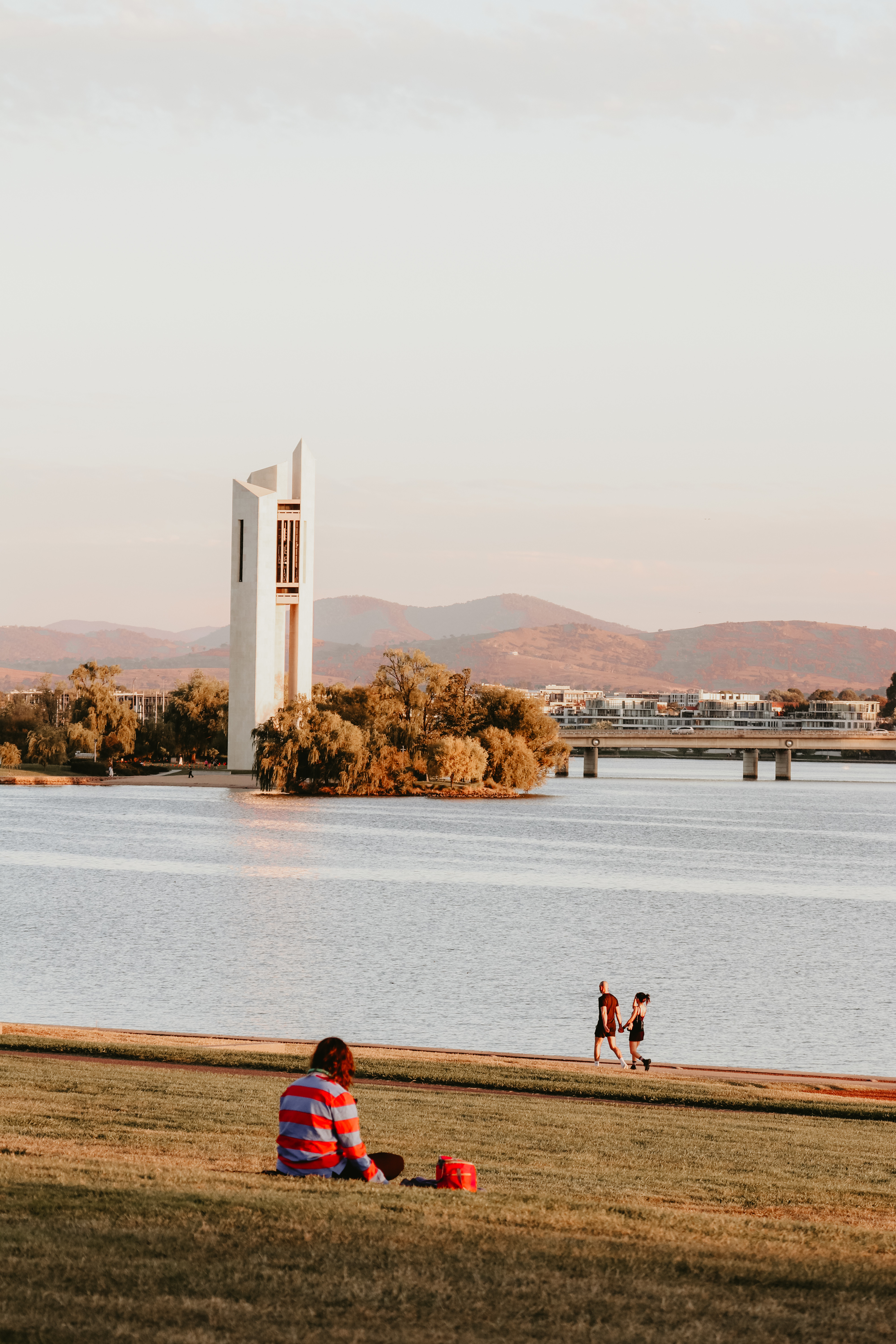 A person sits on a grassy bank by Lake Burley Griffin, with two figures walking near the water's edge. In the background, the National Carillon, a white modern tower, stands on an island near a bridge, with hills in the distance under a pale sky.